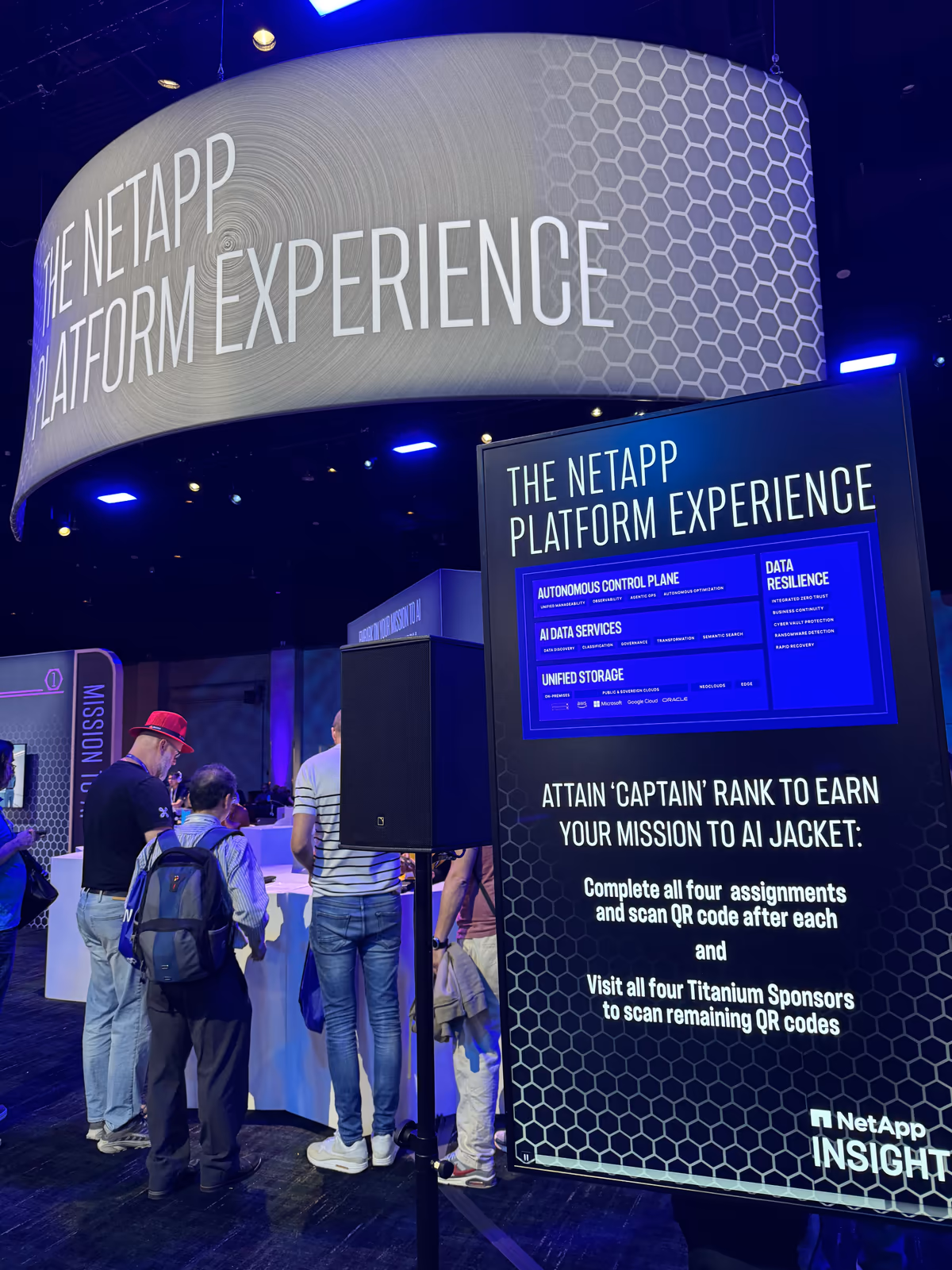 A NetApp INSIGHT sign listing Autonomous Control Plane, AI Data Services, Unified Storage, and Data Resilience, with instructions to complete four Mission to AI assignments to earn a jacket